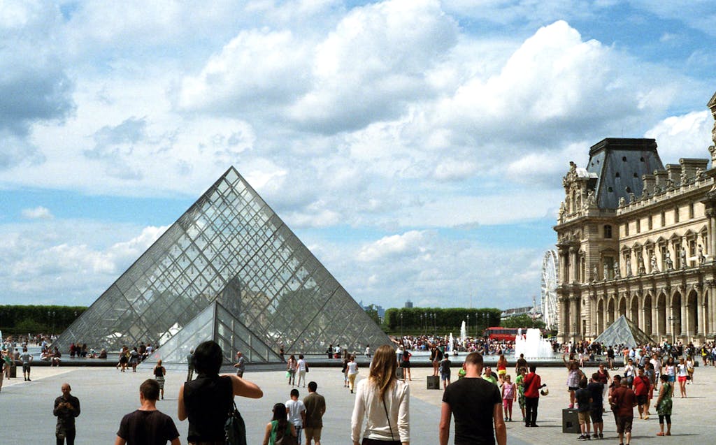 Crowded view of the iconic Louvre Pyramid with tourists in Paris, France on a sunny day.