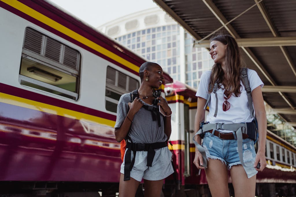 Two women with backpacks smiling and chatting at a train station, ready for urban travel.